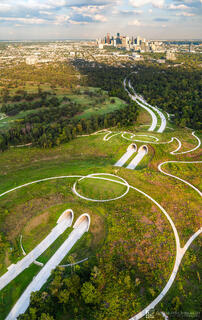 Twin tunnels entering Memorial Park with winding paths and green space leading toward the Houston skyline at sunset, photographed from above.
